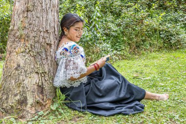 Otavalo, Ekvador: 6 Ağustos 2025: Geleneksel işlemeli giysiler içinde çıplak ayaklı yerli Otavalo kadını, doğal bir And ortamında bir ağaç gövdesine yaslanırken bir kitap okuyor, barış ve kültürel bağlantı kuruyor..