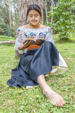 Otavalo, Ekvador: 6 Ağustos 2025: Geleneksel işlemeli giysiler içinde çıplak ayaklı yerli Otavalo kadını, doğal bir And ortamında bir ağaç gövdesine yaslanırken bir kitap okuyor, barış ve kültürel bağlantı kuruyor..