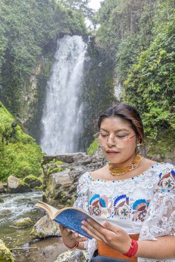 Otavalo, Ekvador: 6 Ağustos 2025: Geleneksel giyinmiş yerli bir Otavalo kadını Peguche şelalesinin yanında okumayı sever. Kültür, bilgelik ve doğayla uyum içinde yaşayan bir sahne..