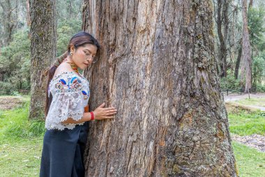 Otavalo, Ekvador: 6 Ağustos 2025: Geleneksel giyinmiş, bir ağaca sarılan yerli Otavalo kadını.