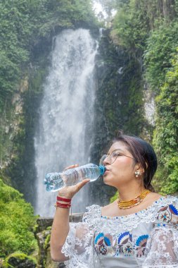 Otavalo, Ekvador: 6 Ağustos 2025: Peguche şelalesinin yanında su şişesi ile su içen geleneksel giyimli yerli bir Otavalo kadını.
