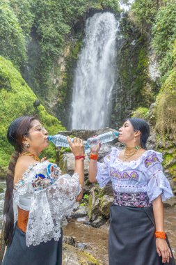 Otavalo, Ekvador: 6 Ağustos 2025: Peguche şelalesinin yanında su şişesi ile su içen geleneksel giyimli yerli bir Otavalo kadını.