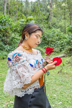 Otavalo, Ekvador: 6 Ağustos 2025: Geleneksel giyinmiş yerli bir Otavalo kadını, doğal bir ortamda kırmızı bir gül kokusunun tadını çıkarıyor, kültürel bağlantı, barış ve ahengi aktarıyor.