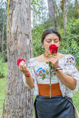 Otavalo, Ekvador: 6 Ağustos 2025: Geleneksel giyinmiş yerli bir Otavalo kadını, doğal bir ortamda kırmızı bir gül kokusunun tadını çıkarıyor, kültürel bağlantı, barış ve ahengi aktarıyor.