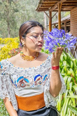 Otavalo, Ecuador - August 6, 2025: Otavalo indigenous woman in traditional attire enjoys the scent of an African agapanthus flower, also known as lily of the Nile, in a natural Andean setting.