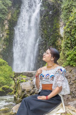 Otavalo, Ecuador - August 6, 2025: Indigenous woman from Otavalo sitting on a rock enjoying the beautiful Peguche waterfall, reflecting Andean tradition, spirituality, and connection with nature.