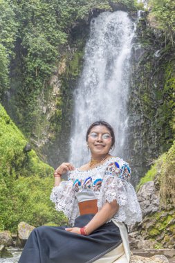 Otavalo, Ecuador - August 6, 2025: Indigenous woman from Otavalo sitting on a rock enjoying the beautiful Peguche waterfall, reflecting Andean tradition, spirituality, and connection with nature.