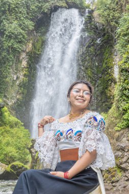 Otavalo, Ecuador - August 6, 2025: Indigenous woman from Otavalo sitting on a rock enjoying the beautiful Peguche waterfall, reflecting Andean tradition, spirituality, and connection with nature.