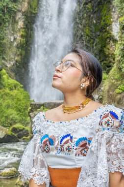 Otavalo, Ecuador - August 6, 2025: Indigenous woman from Otavalo sitting on a rock enjoying the beautiful Peguche waterfall, reflecting Andean tradition, spirituality, and connection with nature.
