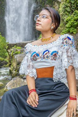 Otavalo, Ecuador - August 6, 2025: Indigenous woman from Otavalo sitting on a rock enjoying the beautiful Peguche waterfall, reflecting Andean tradition, spirituality, and connection with nature.