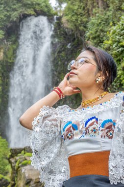 Otavalo, Ecuador - August 6, 2025: Indigenous woman from Otavalo sitting on a rock enjoying the beautiful Peguche waterfall, reflecting Andean tradition, spirituality, and connection with nature.