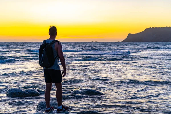 Kumsalda duran sırt çantalı bir adam.