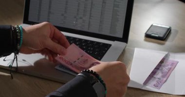 Birisi masada oturup hesap tablolarını gösteren dizüstü bilgisayarına odaklanarak nakit sayıyor. Yakınlarda bir akıllı telefon var, çalışma alanına ekleniyor..