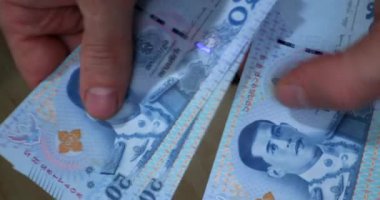 Bir kişi canlı bir piyasa ortamında renkli Tayland banknotlarını sayıp düzenliyor. Satıcılar etraflarındaki potansiyel müşterilere seslenirken güneş batıyor..