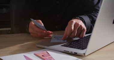 Bireysel hesaplar ve bir dizüstü bilgisayarla ahşap bir masada otururken banknotları idare eder. Çevre loş aydınlık, finansal görevler için odaklanmış bir atmosfer yaratıyor.