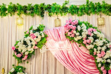 Büyüleyici bir düğün arkaplanı, orta süsü olarak dalgalı pembe bir saten örtü sunar. Canlı yeşil yapraklarla vurgulanmış gür pembe ve beyaz güllerin kemerleri, parlayan neon bir işareti çerçeveler. Üç altın asılı fener bir tutam sıcak tutar..