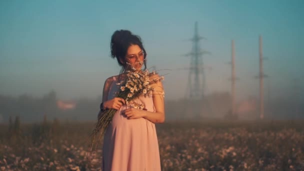 A girl with glasses at dawn holds a bouquet of daisies and strokes her belly. — Stock Video