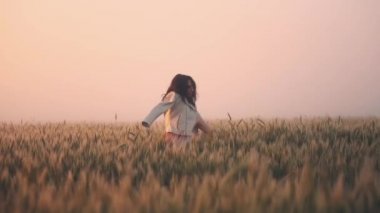 Omzunda ceketi olan hamile bir kız, buğday tarlasında dans ediyor ve dönüyor.