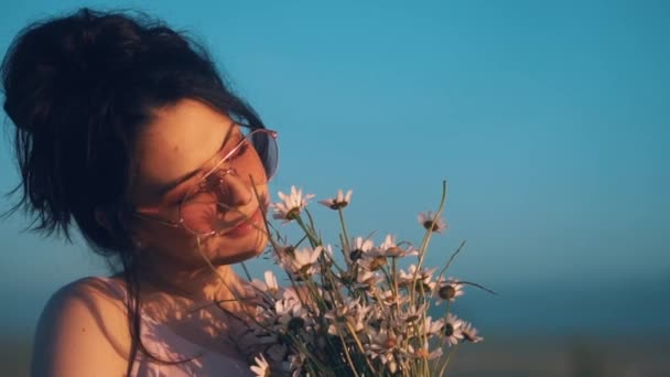 Młoda dziewczyna w różowych okularach patrzy na słońce i wącha bukiet rumianków — Wideo stockowe