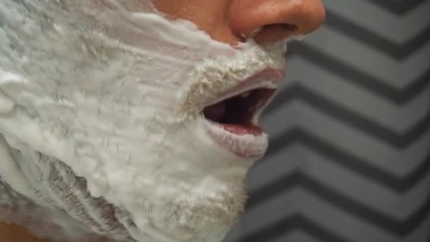 Молодой человек с темными волосами и пеной для бритья на лице открывает рот. — стоковое видео