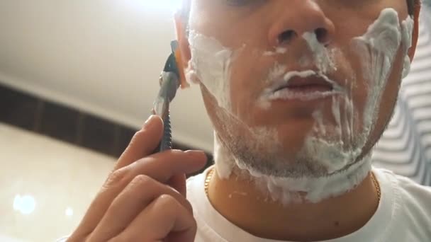 En ung man rakar skäggstubben på kinden med en dålig rakkniv. Skum. Närbild — Stockvideo