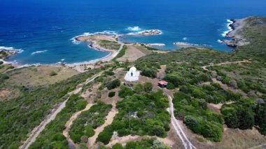 On a Greek island in the Aegean, a white chapel stands against the deep blue Mediterranean sea. The waves reach two coves forming beautiful beaches, while the coastline is surrounded by dense green vegetation.
