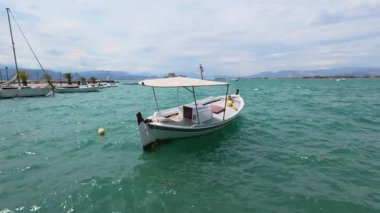 A traditional Greek port shows a wooden fishing boat moving in the Mediterranean waters, with ships and a castle visible in the background.