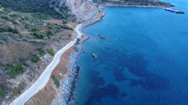 A Greek coastal roadway winds along the blue Mediterranean Sea, ending at a small harbor protected by cliffs from the open waters.
