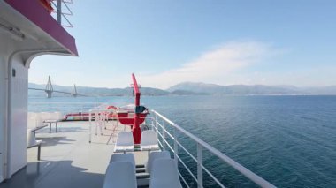 A ship sails through the Rio Antirrio passage in Greece. The famous suspension bridge is visible with a red lifeboat in the foreground.