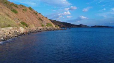 A coastal road in Greece runs beside the Mediterranean Sea on a sunny summer day. Waves crash against the rocks just meters from the road, which winds through steep cliffs and rugged mountains.
