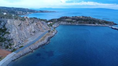 A coastal road in Greece runs beside the Mediterranean Sea at sunset. Waves strike the rocks near the asphalt, while the road leads to a picturesque harbor.