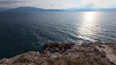 At sunset, the Aegean Sea glows as waves strike a rocky coast in Greece. Distant islands appear under golden Mediterranean light.