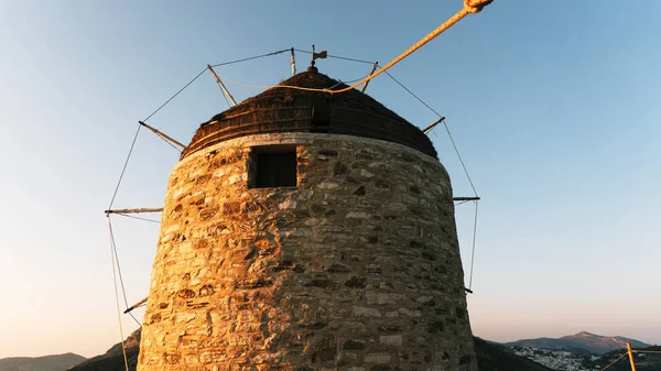 An old stone mill on top of a mountain, against the backdrop of a mountain range and valley with a small town with white houses on a Greek island