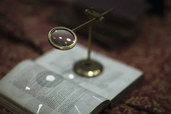 Vintage loupe close-up above a book among vintage items on a table on a red tablecloth with a gold pattern