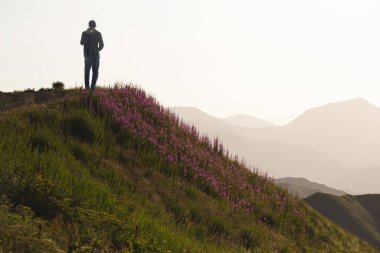 Sırt çantalı genç bir turist, şafak vakti dağlarda çiçek açan yamaçların arkasında gün batımına hayran. Güneş ışığının sisi ile uzak dağ zincirleri