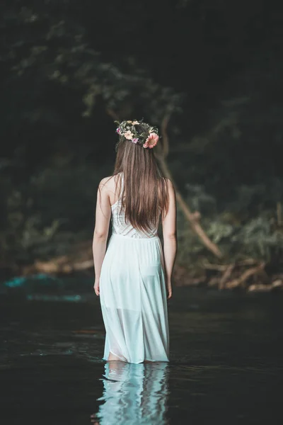 Fine art outdoor portrait of beautiful young woman in a white dress and floral wreath. Girl walking alone by the river