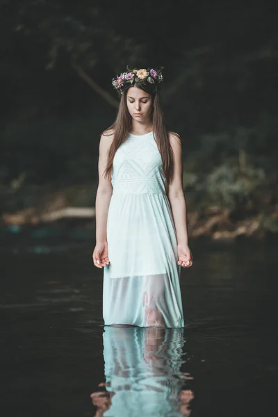 Fine art outdoor portrait of beautiful young woman in a white dress and floral wreath. Girl walking alone by the river