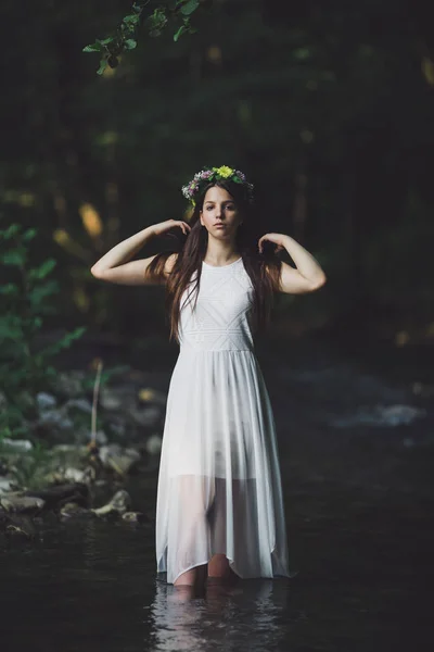 Fine art outdoor portrait of beautiful young woman in a white dress and floral wreath. Girl walking alone by the river