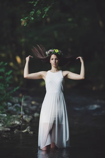 Fine art outdoor portrait of beautiful young woman in a white dress and floral wreath. Girl walking alone by the river