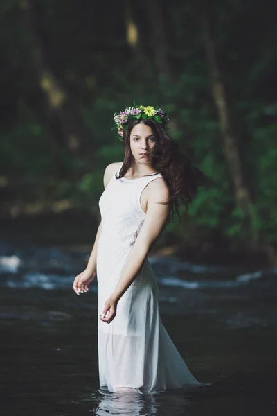 Fine art outdoor portrait of beautiful young woman in a white dress and floral wreath. Girl walking alone by the river