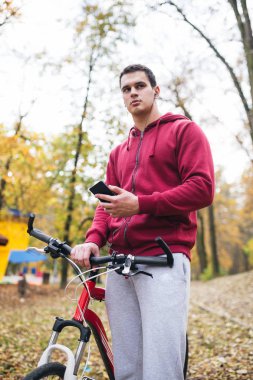 Sonbahar açık havada. Bisiklet Park ile ayakta, bir cep telefonu tutan ve müzik toplama kulaklık genç ciddi yakışıklı adamla izler.