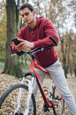 Sonbahar açık havada. Bisiklet Park ile ayakta, bir cep telefonu tutan ve müzik toplama kulaklık genç ciddi yakışıklı adamla izler.
