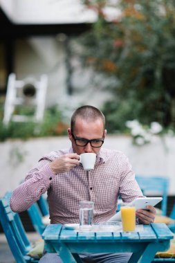 Kafe barda oturmuş, kahve içme ve tablet kullanarak gözlük ile raslantı giyimli adam.