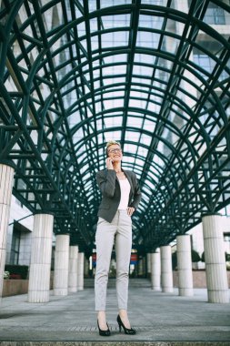 Mutlu ve kendine güvenen genç iş kadını büyük modern binası önünde ayakta. O gülümseyen ve onu cep telefonundan konuşuyor. 