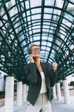 Mutlu ve kendine güvenen genç iş kadını büyük modern binası önünde ayakta. O gülümseyen ve onu cep telefonundan konuşuyor. 