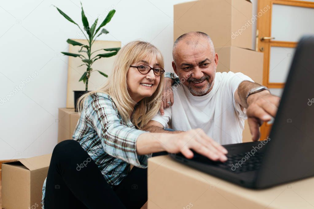 Middle aged couple sitting between boxes in their new apartment and using a laptop for some interior decorating ideas. Real estate theme.