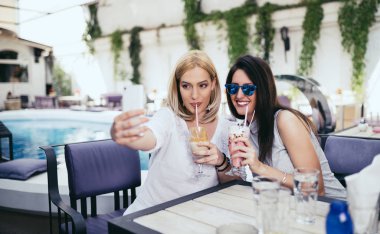 İki genç ve güzel kadın özel kokteyl evi partide zevk. Oturuyorlar, gülümsüyorlar ve selfie fotoğrafı çekiyorlar. Arka planda yüzme havuzu.