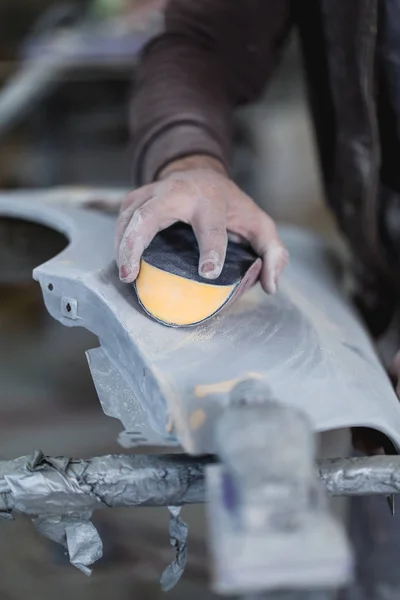 Car detailing - Man with sandpaper in auto repair shop sanding polishing and preparing car parts for painting. Selective focus on man\'s hand.