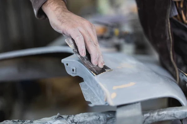 Car detailing - Man with sandpaper in auto repair shop sanding polishing and preparing car parts for painting. Selective focus on man\'s hand.