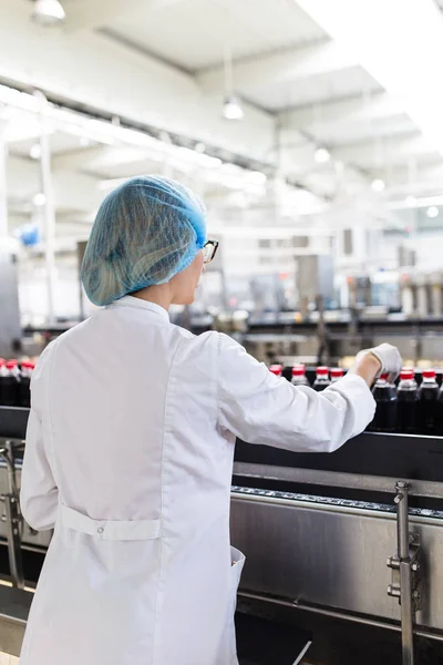 严重的中年妇女质量控制工作者检查机器人线装瓶和包装碳酸黑汁的软饮料瓶 — 图库照片